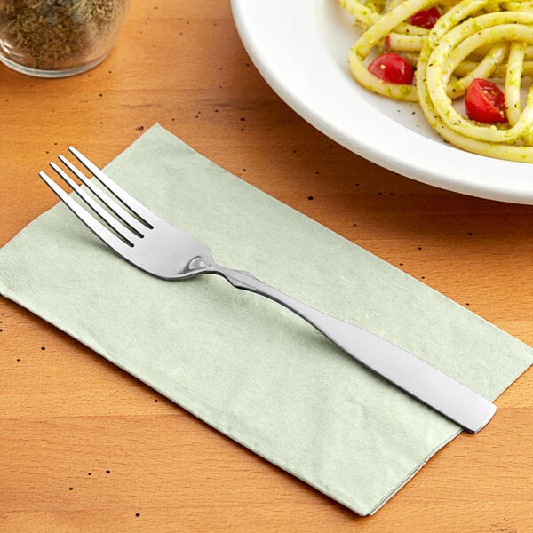 A Choice stainless steel dinner fork on a napkin next to a plate of spaghetti.