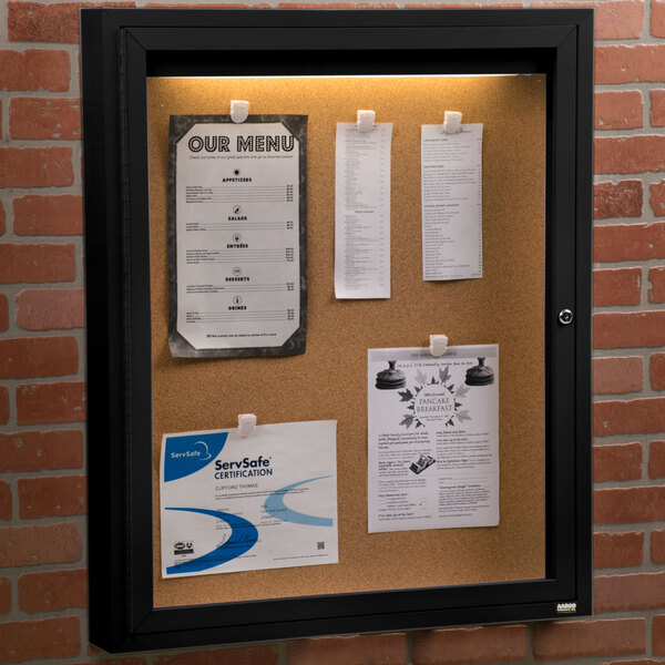 An Aarco black indoor lighted bulletin board cabinet with cork papers pinned to it.
