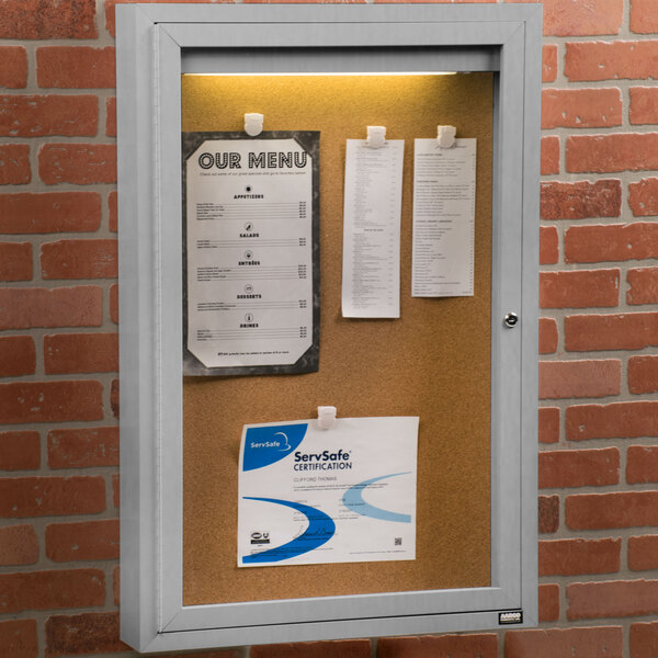 An Aarco satin anodized indoor lighted bulletin board cabinet with a white cork board and paper on it.