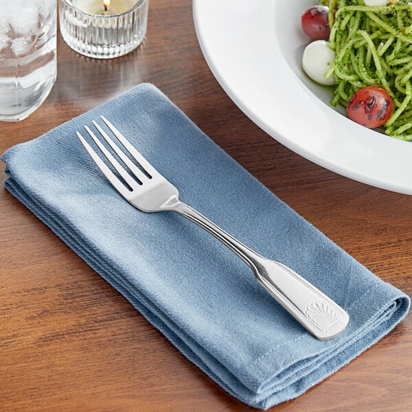 An Acopa stainless steel dinner fork on a napkin next to a plate of food.
