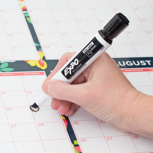 A hand using a black Expo chisel tip dry erase marker to write on a calendar.