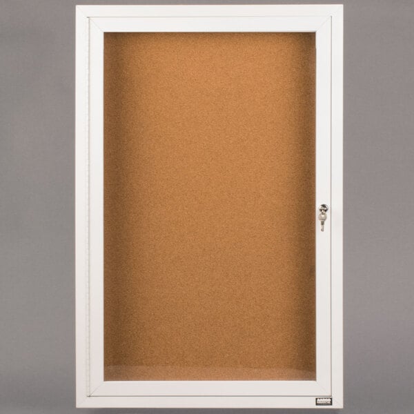 A white cabinet with a glass door and key enclosing a cork bulletin board.