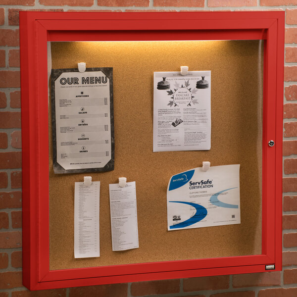 An Aarco red indoor bulletin board cabinet with papers attached to it.