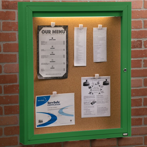 An Aarco green indoor lighted bulletin board cabinet with cork board inside and papers attached to it.