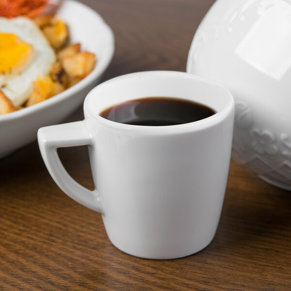 A Schonwald white porcelain espresso cup filled with coffee on a saucer next to a plate of food.