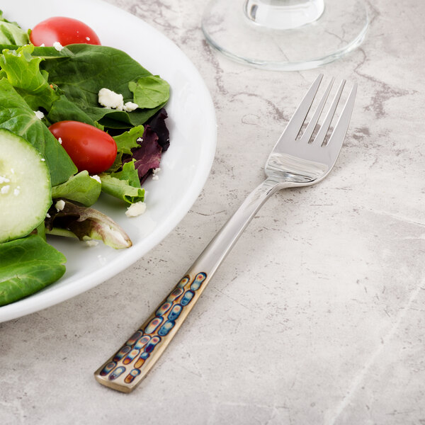 A Master's Gauge stainless steel salad fork next to a plate of salad with tomatoes and lettuce.