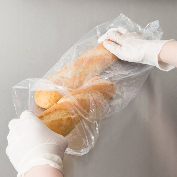 A pair of gloved hands holding a plastic bag of bread with a baguette inside.