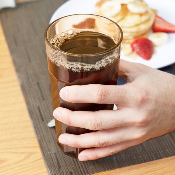 A hand holding a Cambro amber plastic tumbler of brown liquid.