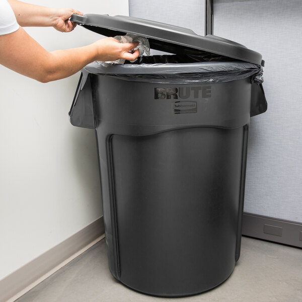 A woman's hands putting a plastic bag into a black Rubbermaid BRUTE trash can in a corporate office cafeteria.