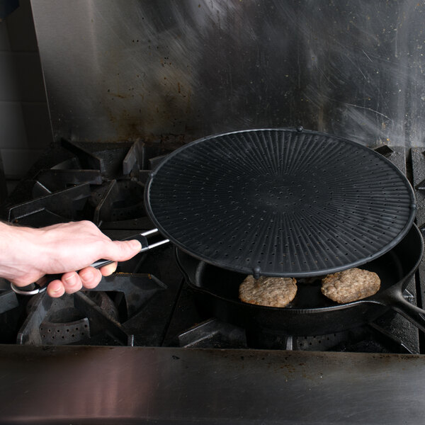 A hand holding a black Mastrad silicone splatter screen over food cooking in a pan.