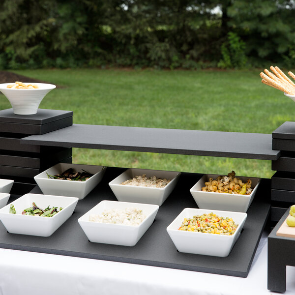 A black wood Vollrath display shelf holding white square bowls of food on a black surface.