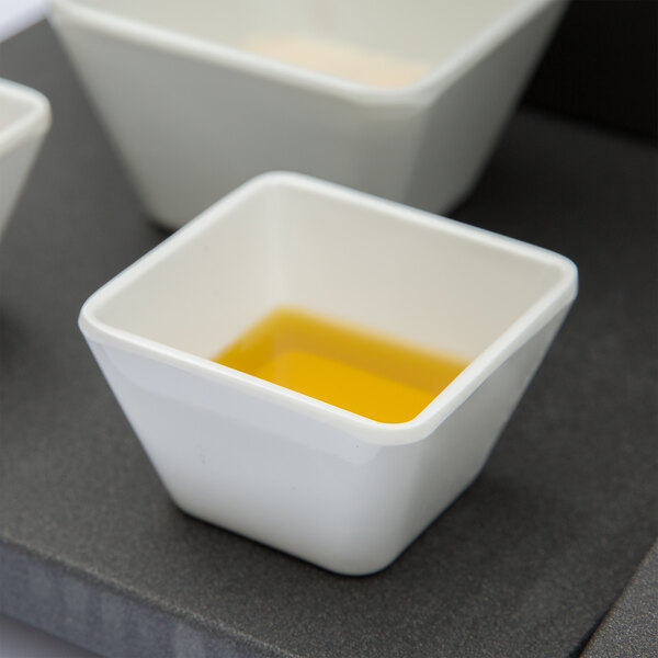 A white square Vollrath melamine bowl with yellow liquid inside.