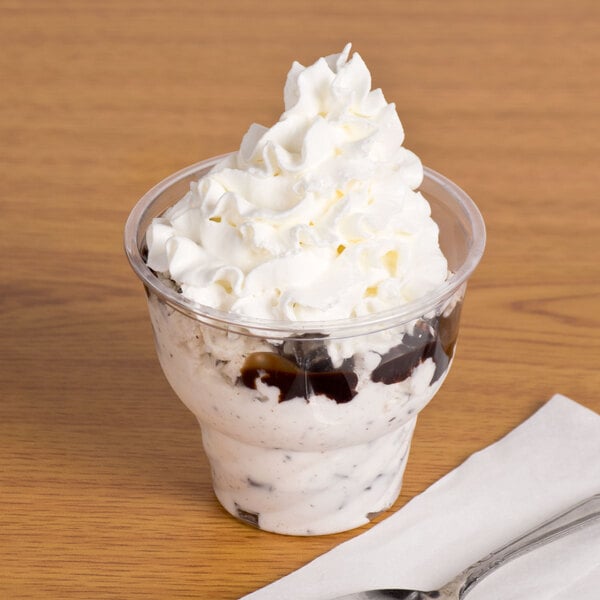 A Fabri-Kal clear plastic sundae cup filled with ice cream, whipped cream, and chocolate syrup with a spoon.