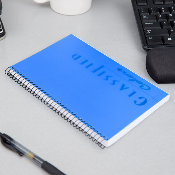 A white wirebound notebook with indigo cover and black spiral on a desk with a pen.