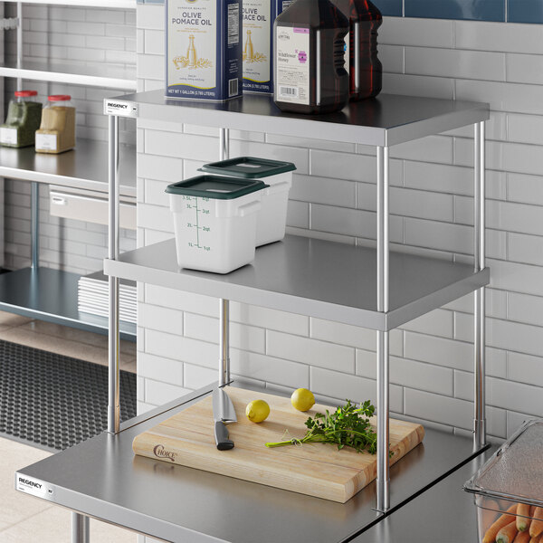 A stainless steel double-deck overshelf with two tiers, holding containers and bottles, positioned above a countertop with a cutting board, knife, lemons, and herbs.