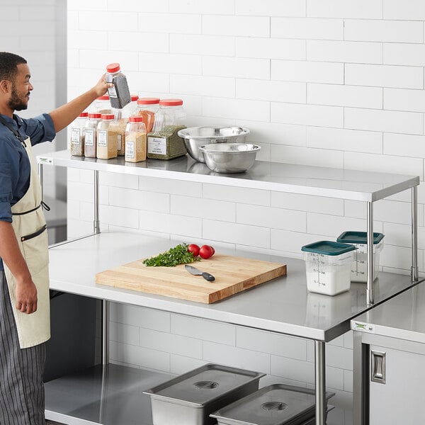 A man in a professional kitchen standing at a counter with a Regency stainless steel table mounted overshelf.