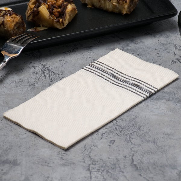 A Hoffmaster white dishtowel print dinner napkin with black stripes next to a fork on a table.