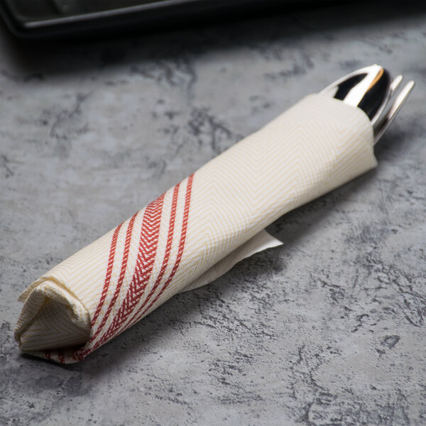 A white napkin with a red dishtowel print wrapped around silverware on a table.