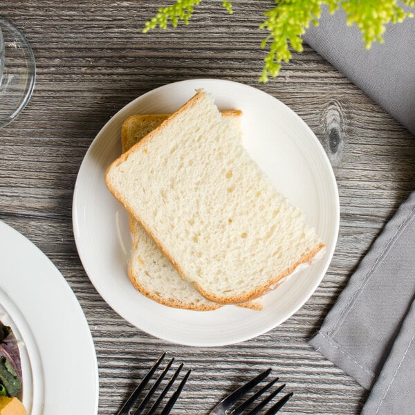 A Bon Chef white porcelain bread plate with bread and a fork on a table with salad.