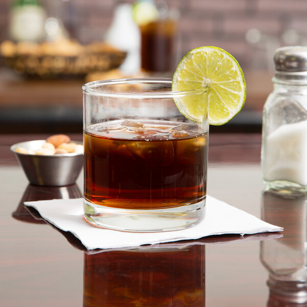 A Libbey rocks glass of brown liquid with ice and a lime wedge.