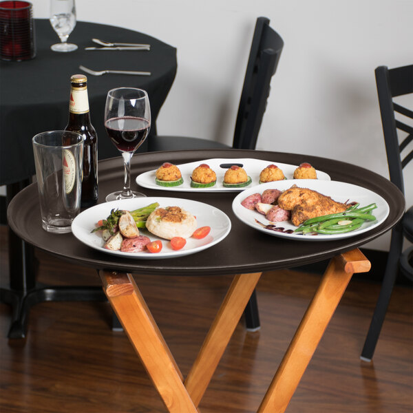 A wooden table with a Cambro Tavern Tan non-skid oval serving tray holding plates of food and glasses of wine.