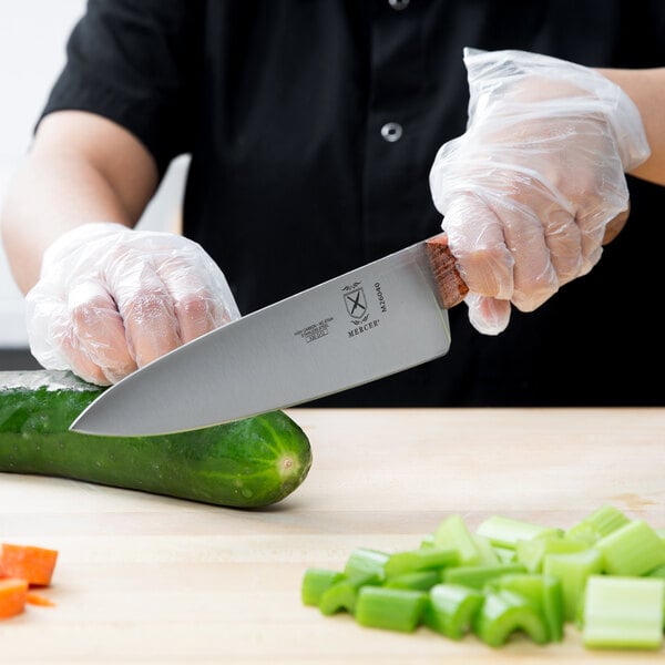 A person in gloves uses a Mercer Praxis chef's knife to cut a cucumber on a cutting board.