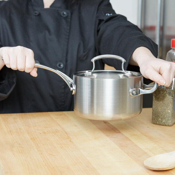 A woman in a chef's uniform holding a Vollrath stainless steel sauce pan over a wooden surface.