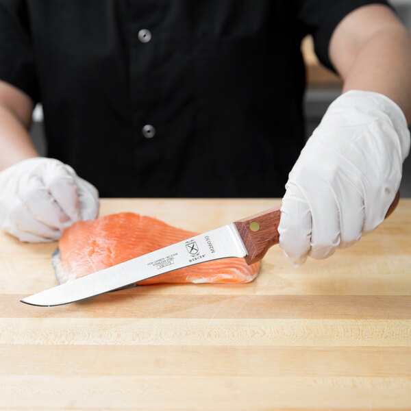 A person in white gloves using a Mercer Culinary Praxis Stiff Boning Knife to cut a piece of salmon on a cutting board.