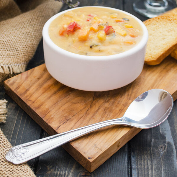 A bowl of soup on a wood board next to a Walco Danish Pride stainless steel bouillon spoon.