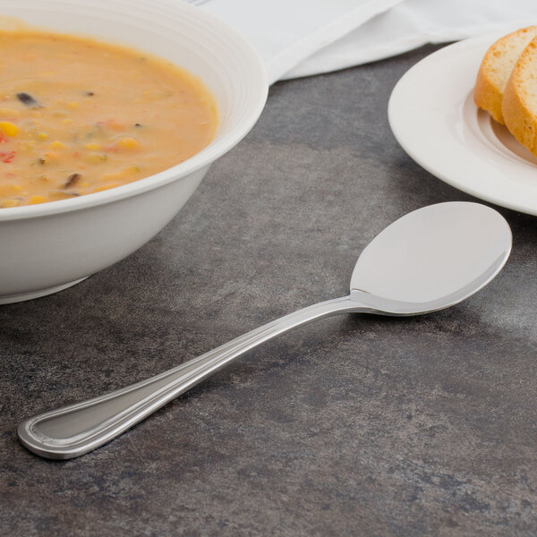 A bowl of soup with a Walco Lisbon bouillon spoon next to a plate of bread on a table.