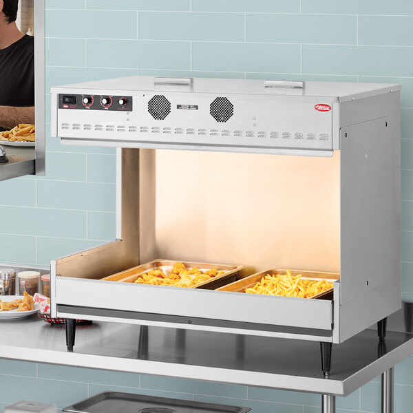 A Hatco multi-product warming station on a stainless steel counter over an oven with food in trays.