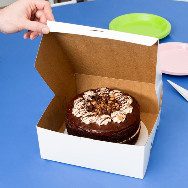 A hand holding a 10" x 10" white cake box with a chocolate cake inside.