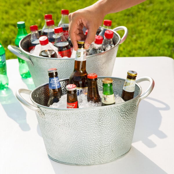 A hand holding a bottle of beer in a Tablecraft galvanized steel beverage bucket on a table.