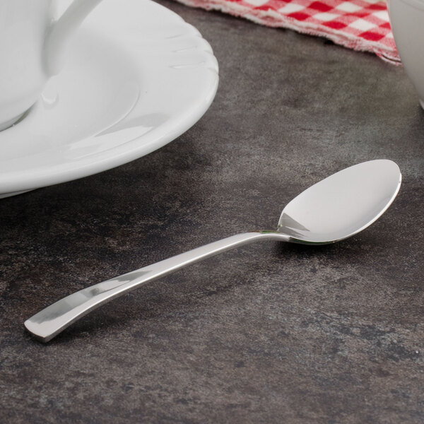 A Walco Freya stainless steel demitasse spoon on a table next to a white cup with a red and white checkered cloth.
