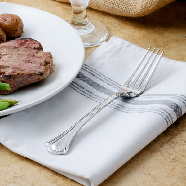 A Walco stainless steel dinner fork on a napkin next to a plate of meat and vegetables.