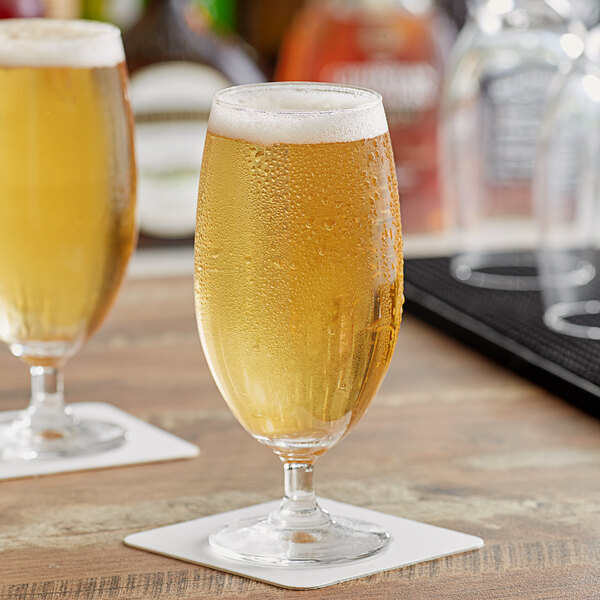 Two Acopa stemmed pilsner glasses of beer on a table with a bottle of liquor.