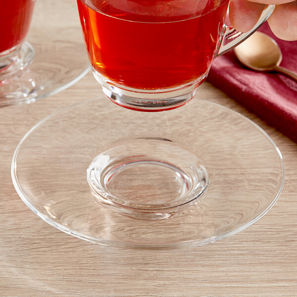 A hand holding a glass Acopa coffee saucer with a cup of red liquid on a red tablecloth.