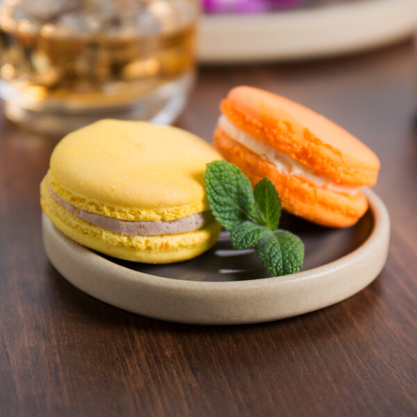 A Chef & Sommelier gray stoneware plate with two macarons, one yellow and one brown, and a mint leaf on a table.