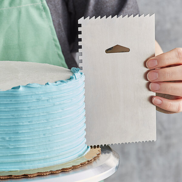 A hand holding a white rectangular Ateco Decorating and Icing Comb over a blue and white decorated cake.