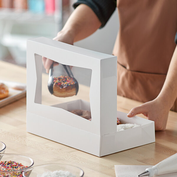 A person putting a doughnut into a white Baker's Mark bakery box with a window.