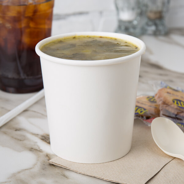 A white Choice paper food cup filled with soup on a table with a spoon.