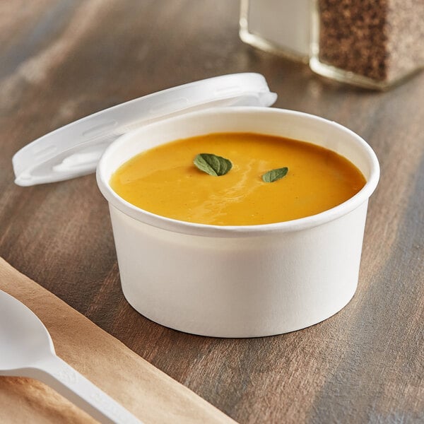 A close up of a bowl of soup in a white paper cup with a vented plastic lid and a white plastic spoon.