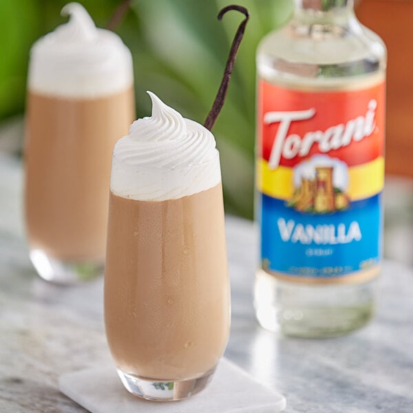 A glass of brown liquid with whipped cream on top and a bottle of Torani Vanilla Flavoring Syrup.