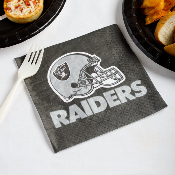 A Creative Converting Las Vegas Raiders luncheon napkin with the Raiders logo on it on a table with a plate of food.