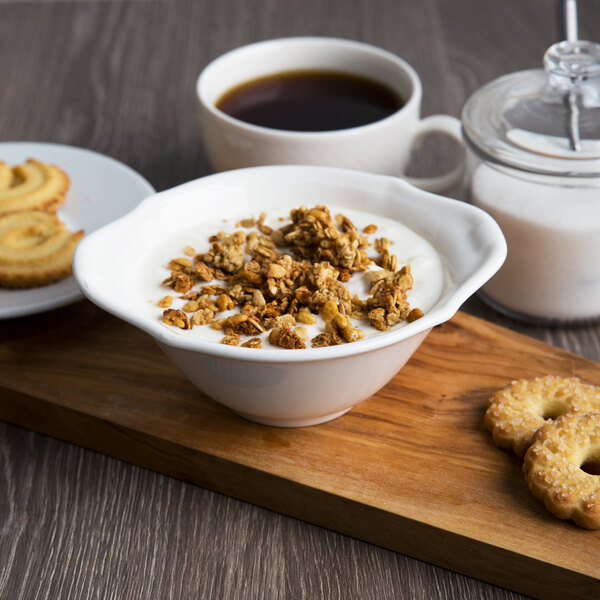 A white porcelain soup cup filled with granola and cookies on a wood board.