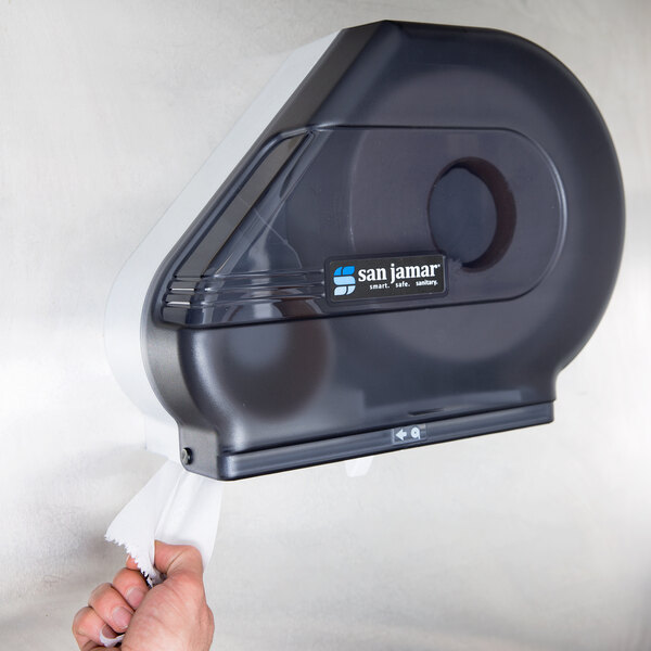 A hand pulling tissue paper from a black San Jamar toilet paper dispenser.