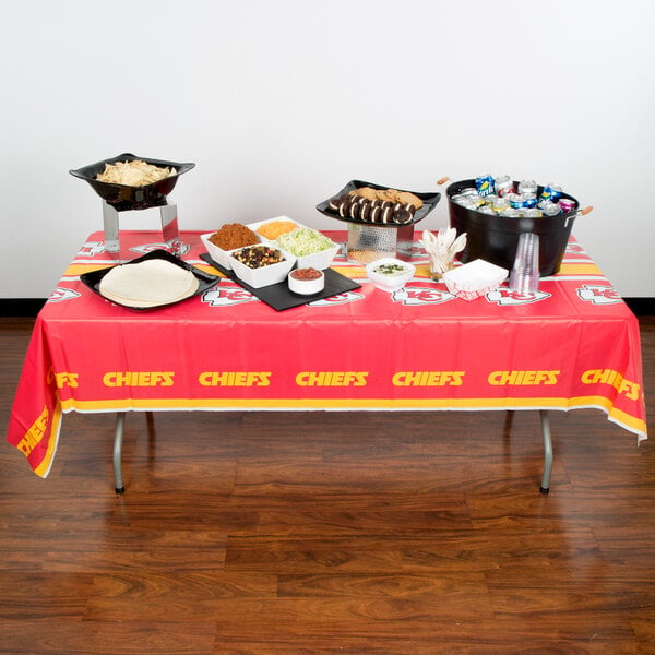 A Kansas City Chiefs plastic table cover on a table with food and drinks on it.