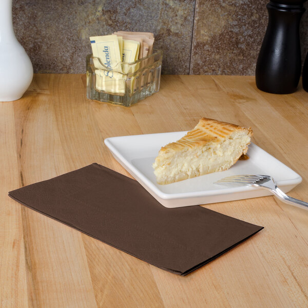 A plate with a slice of cheesecake and a fork on a chocolate brown Hoffmaster paper napkin.