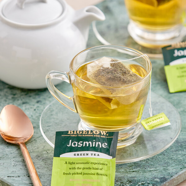 A glass cup of Bigelow Jasmine Green Tea with a tea bag in it on a table.