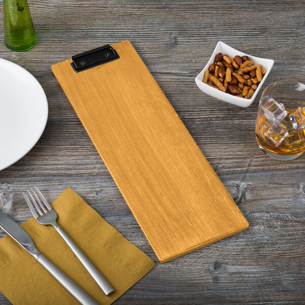 A customizable oak wood menu clipboard on a table with a plate and glass of ice with a fork and knife on a napkin.